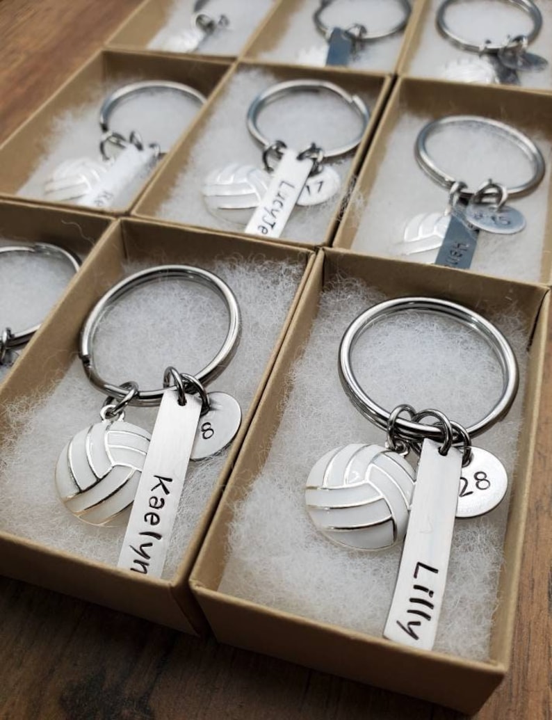 May include: A set of personalized volleyball keychain gifts in small brown boxes. Each keychain features a silver volleyball charm, a silver tag with a name, and a silver charm with a number. The names on the keychains are Kaelyn, Lilly, and Lucy. The numbers on the keychains are 17, 28, and 8.