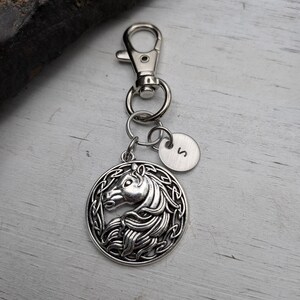 May include: Silver-toned keychain featuring a round pendant with a horse head design. The pendant is attached to a clasp and a small circular charm with the letter "S". The horse head is surrounded by an intricate border.
