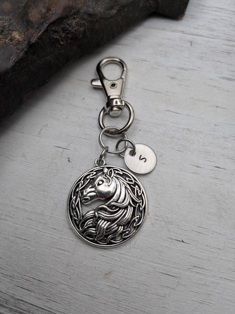 May include: Silver-toned keychain featuring a round pendant with a horse head design. The pendant is attached to a clasp and a small circular charm with the letter "S". The horse head is surrounded by an intricate border.