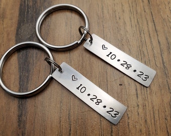 May include: Two silver keychains with a heart and the date 10.28.23 engraved on each. The keychains are on a wooden surface.
