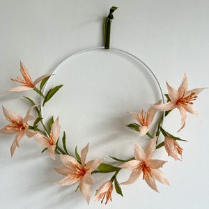May include: A peach-colored paper flower wreath with green leaves hangs on a white wall. The wreath is made of delicate paper flowers and is attached to a white metal hoop.