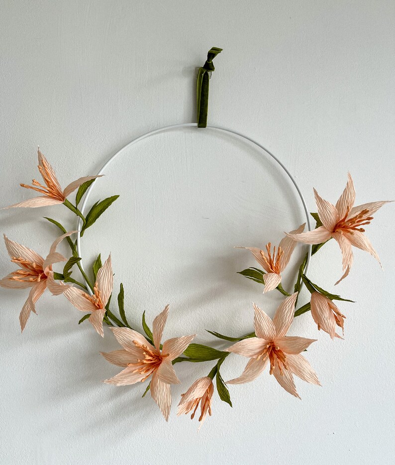 May include: A peach-colored paper flower wreath with green leaves hangs on a white wall. The wreath is made of delicate paper flowers and is attached to a white metal hoop.