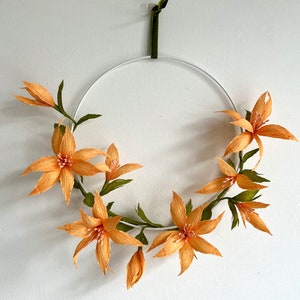 May include: A white metal wreath with a green ribbon hanging from the top. The wreath is decorated with orange paper flowers.