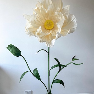 May include: A large, white paper flower with a yellow center. The flower is on a green stem with green leaves. The flower is standing on a green base.