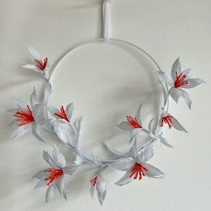 May include: White paper flower wreath with red centers. The wreath is hanging from a white string.