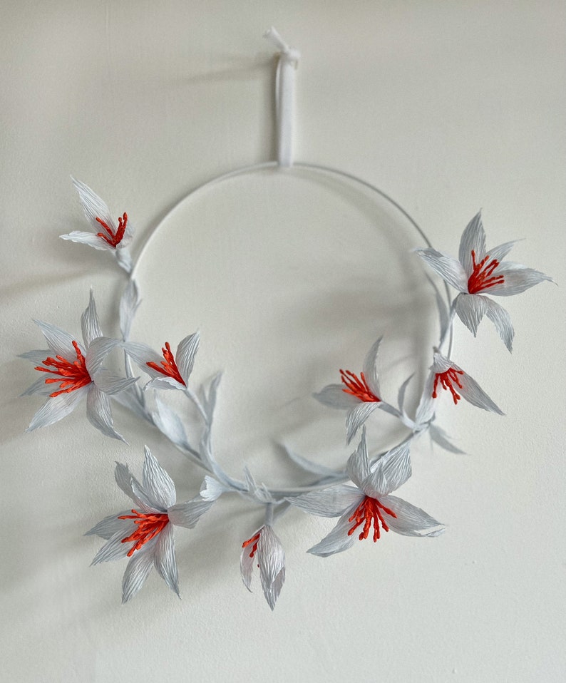 May include: White paper flower wreath with red centers. The wreath is hanging from a white string.