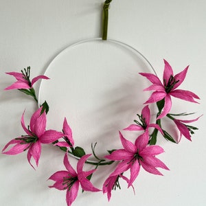 May include: A pink paper flower wreath hanging on a white wall. The wreath is made of several pink paper flowers with green stems and leaves.