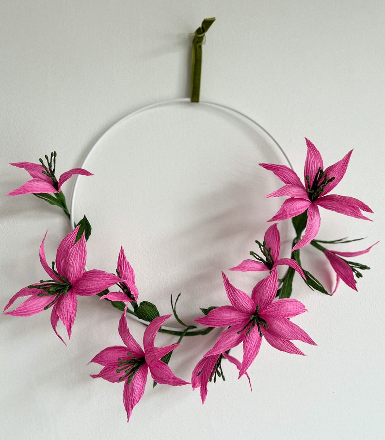 May include: A pink paper flower wreath hanging on a white wall. The wreath is made of several pink paper flowers with green stems and leaves.