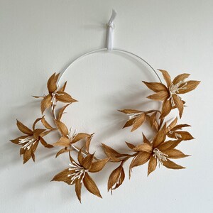 May include: A brown paper flower wreath with white accents hangs on a white wall. The wreath is made of paper flowers and is attached to a white metal hoop.
