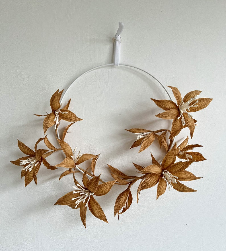May include: A brown paper flower wreath with white accents hangs on a white wall. The wreath is made of paper flowers and is attached to a white metal hoop.
