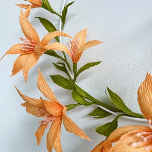 May include: A wreath of peach-colored paper flowers with green paper leaves. The flowers are arranged in a circle around a white wire frame.