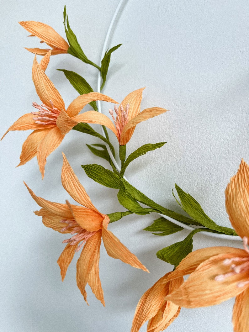 May include: A wreath of peach-colored paper flowers with green paper leaves. The flowers are arranged in a circle around a white wire frame.