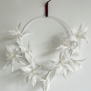 May include: A white paper flower wreath with a burgundy ribbon hanging from the top. The wreath is made of several white paper flowers with a delicate, crinkled texture.