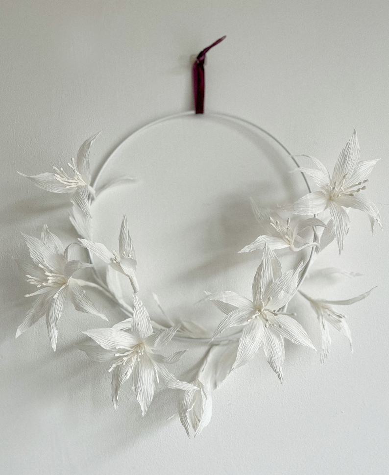 May include: A white paper flower wreath with a burgundy ribbon hanging from the top. The wreath is made of several white paper flowers with a delicate, crinkled texture.