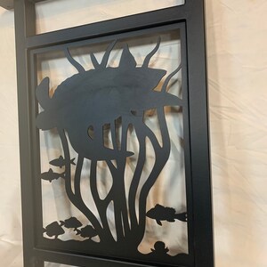 May include: Black metal wall decor featuring a cutout design of a sea turtle swimming through seaweed with fish. The design is framed by a black metal square.