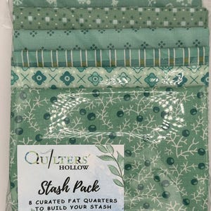 Può includere: Una collezione di otto fat quarters in tonalità di verde, confezionati in plastica trasparente. I tessuti presentano vari motivi, tra cui fiocchi di neve, treni e disegni floreali. La confezione include un'etichetta con "Quilters' Hollow Stash Pack", dimensioni di circa 45.7cm x 55.9cm.