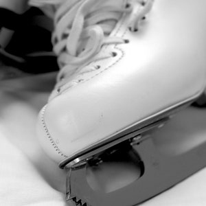 May include: A close-up shot of a white ice skate with laces. The skate features a curved blade with a toothed edge. The boot is made of a smooth material and has visible stitching. The image is in monochrome.