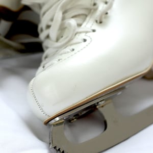 May include: Close-up of a white ice skate with laces and a silver blade. The skate is made of leather and has a classic design. The blade has a serrated edge. The skate is on a white surface.