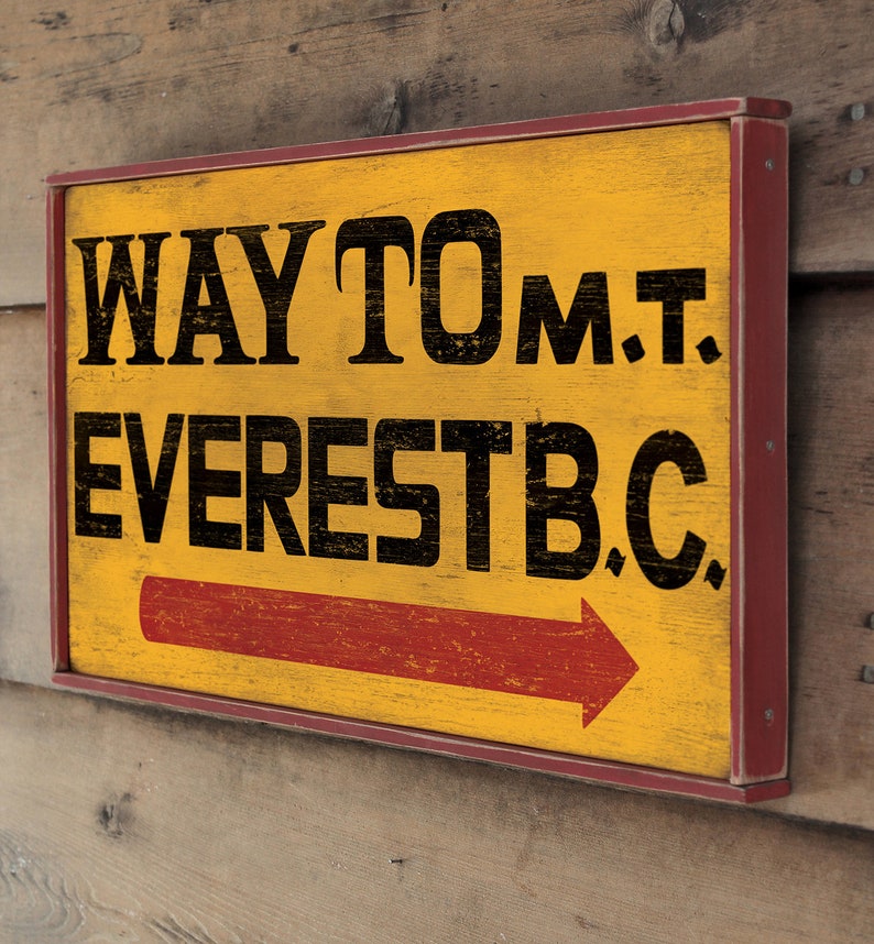 May include: A yellow wooden sign with red trim and black lettering that reads "WAY TO MT. EVEREST B.C." with a red arrow pointing to the right.