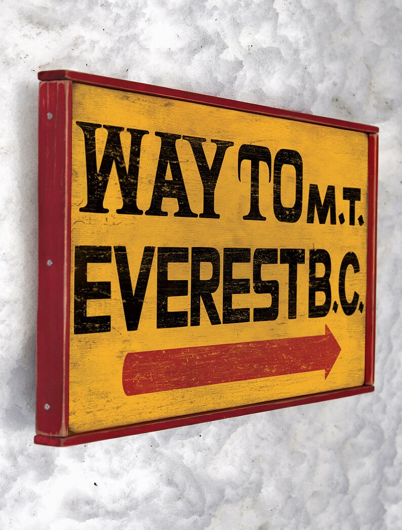 May include: A yellow wooden sign with red trim and a red arrow pointing to the right. The sign reads "WAY TO MT. EVEREST B.C."