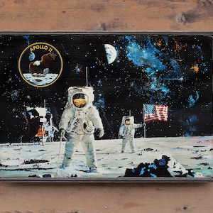 May include: A framed print of a vintage illustration depicting two astronauts walking on the moon. The astronauts are wearing white spacesuits and helmets. The moon's surface is gray and rocky. The background is a dark blue sky with stars and a galaxy. The print includes the text "Apollo II" and a small image of an eagle.