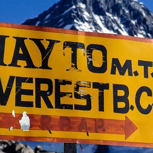 May include: A yellow sign with black text that reads "WAY TO M.T. EVEREST B.C." with a red arrow pointing to the right. The sign is in front of a snow-capped mountain.