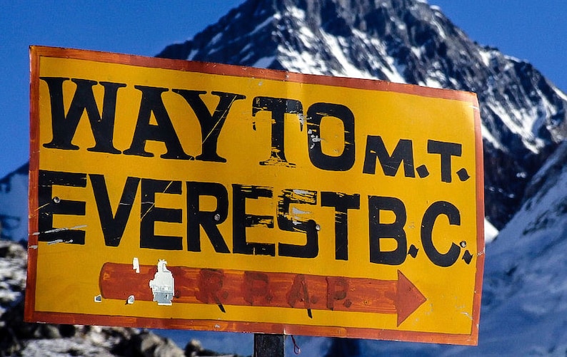 May include: A yellow sign with black text that reads "WAY TO M.T. EVEREST B.C." with a red arrow pointing to the right. The sign is in front of a snow-capped mountain.