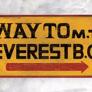 May include: A yellow wooden sign with red trim and a red arrow pointing to the right. The sign reads "WAY TO MT. EVEREST B.C."