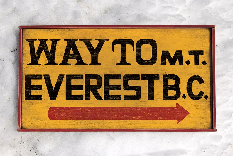 May include: A yellow wooden sign with red trim and a red arrow pointing to the right. The sign reads "WAY TO MT. EVEREST B.C."