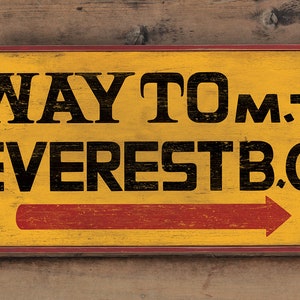 May include: A yellow wooden sign with red trim and a red arrow pointing to the right. The sign reads "WAY TO MT. EVEREST B.C."