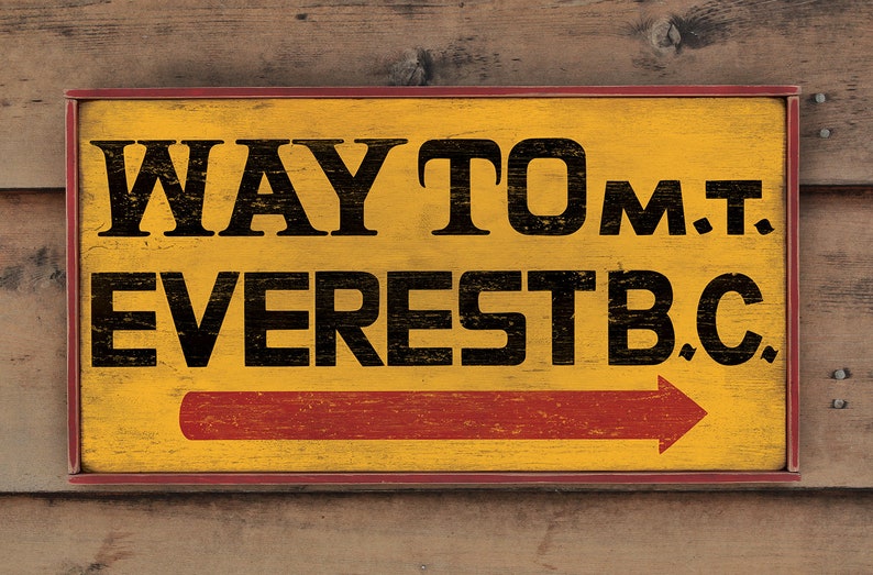 May include: A yellow wooden sign with red trim and a red arrow pointing to the right. The sign reads "WAY TO MT. EVEREST B.C."