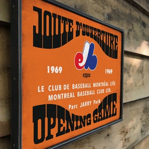 May include: A vintage-style wooden sign with a black frame and orange background. The sign reads "JOUTE D'OUVERTURE" in a retro font, with the Montreal Expos logo and the years 1969. The text below reads "LE CLUB DE BASEBALL MONTR&Eacute;AL LT&Eacute;E MONTREAL BASEBALL CLUB LTD. Parc JARRY Park OPENING GAME".