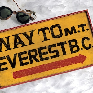 May include: A yellow wooden sign with red trim and a red arrow pointing right. The sign reads "WAY TO M.T. EVEREST B.C."