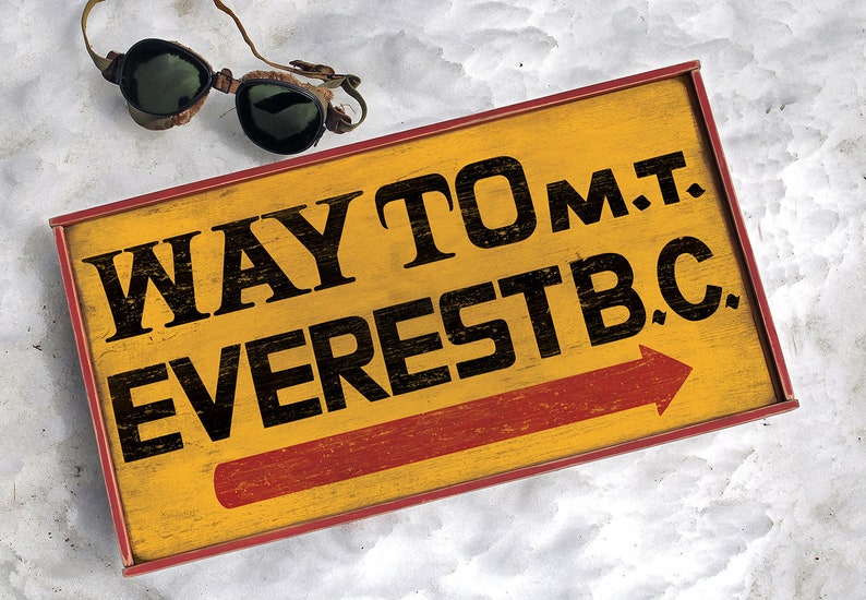 May include: A yellow wooden sign with red trim and a red arrow pointing right. The sign reads "WAY TO M.T. EVEREST B.C."