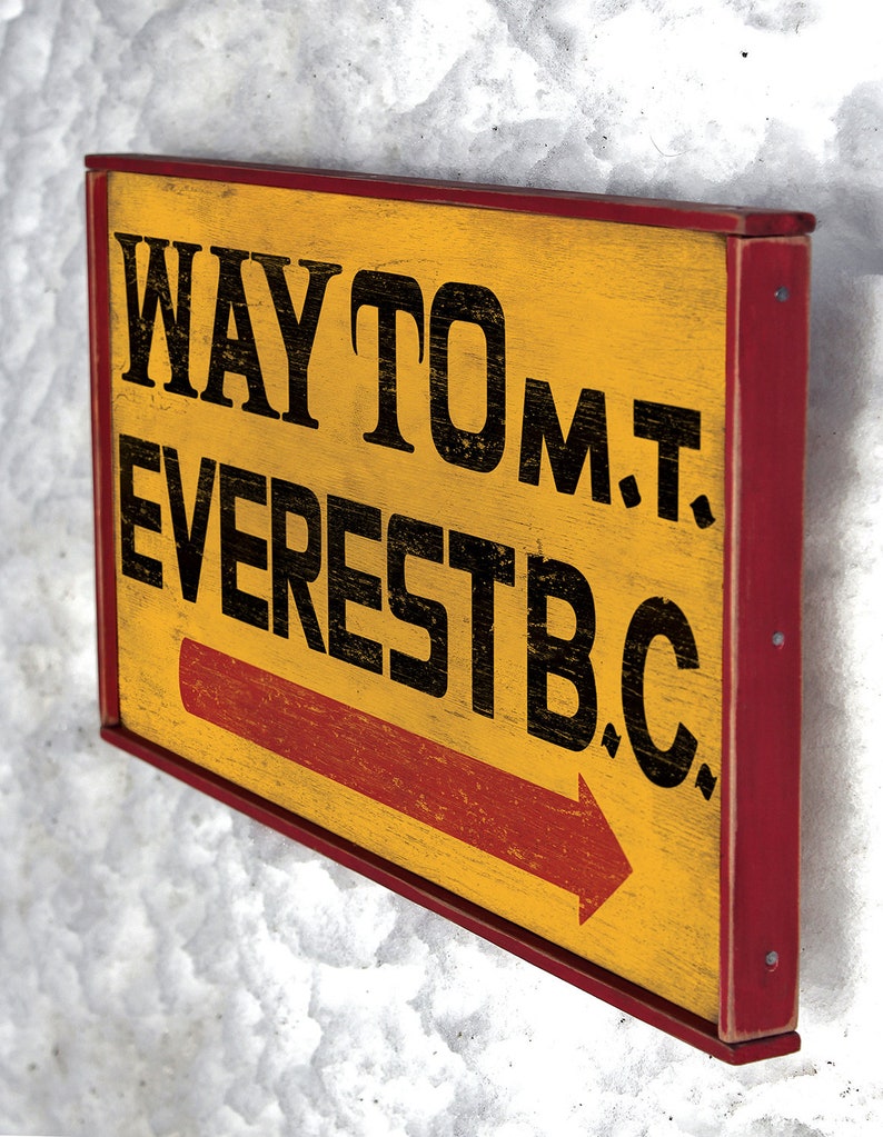 May include: A yellow wooden sign with red trim and a red arrow pointing to the right. The sign reads "WAY TO MT. EVEREST B.C."