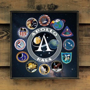 May include: Framed artwork showcasing Apollo mission patches. The central design features the Apollo logo with a planet and a moon. Various mission patches surround the logo, set against a starry background. The frame is dark.