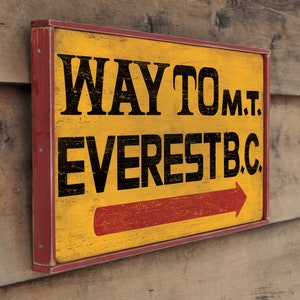 May include: A yellow wooden sign with red trim and black lettering that reads "WAY TO MT. EVEREST B.C." with a red arrow pointing to the right.