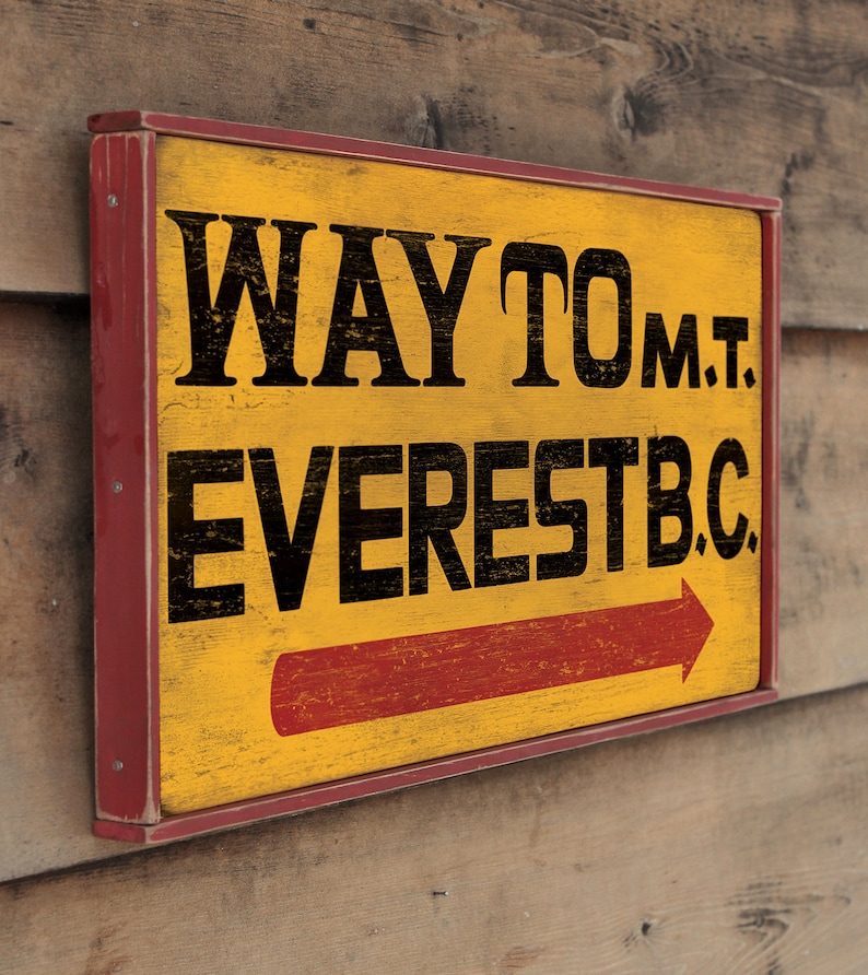 May include: A yellow wooden sign with red trim and black lettering that reads "WAY TO MT. EVEREST B.C." with a red arrow pointing to the right.