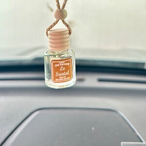 May include: A clear glass bottle with a wooden cap and a gold string hanging from it. The bottle is labeled "Luxury Car Cologne Le Santal" and "Made by Benjamin Murphy".