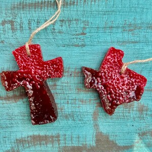 May include: Two red, textured ornaments: a cross and the state of Texas. Each has a twine loop for hanging. The ornaments are set against a weathered, turquoise-colored wooden surface.