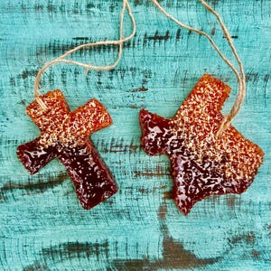 May include: Two brown and orange wax melts, one shaped like a cross and the other shaped like the state of Texas, are hanging from twine on a blue wooden background.