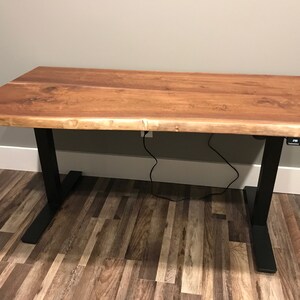 K&ouml;nnte beinhalten: Ein Stehpult mit einer nat&uuml;rlichen Holzplatte und schwarzen Metallbeinen. Der Schreibtisch hat ein Live-Edge-Design und ein Bedienfeld zum Einstellen der H&ouml;he.