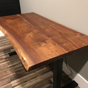 K&ouml;nnte beinhalten: Ein dunkelbrauner Holzschreibtisch mit einer Live-Edge-Oberfl&auml;che und schwarzen, verstellbaren Metallbeinen. Der Schreibtisch steht in einem Raum mit Holzboden und einer hellgrauen Wand.