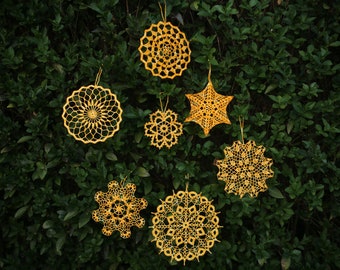 Puede incluir: Siete adornos de ganchillo amarillos colgando de un arbusto verde. Los adornos tienen forma de estrellas, copos de nieve y círculos.