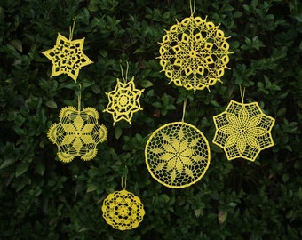 Puede incluir: Siete doilies de ganchillo amarillos colgando de un arbusto verde. Los doilies son de varias formas, incluyendo estrellas, círculos y flores.