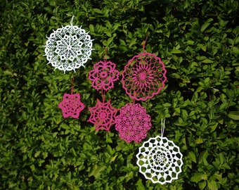 Puede incluir: Una colección de siete doilies de ganchillo colgados de un arbusto verde. Los doilies son de dos colores: blanco y rosa. Los doilies son de diferentes tamaños y formas.