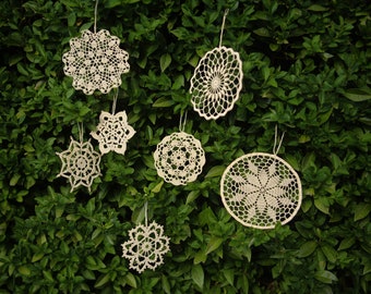 Puede incluir: Siete doilies de ganchillo blancos colgando de un arbusto verde. Los doilies son de diferentes tamaños y formas, algunos con intrincados diseños florales.
