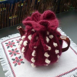 以下が含まれることがあります： 白い水玉模様のマルーン色のティーコージーが、茶色のティーポットを覆っています。ティーコージーは、赤と黒の花柄とフリンジが付いた白い刺繍のプレースマットの上にあります。シーンは濃い緑色のテーブルに設定されています。