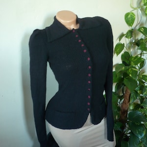 May include: A black knitted cardigan with a collared neckline and long sleeves. The cardigan has burgundy buttons and two small pockets. The garment is displayed on a mannequin.