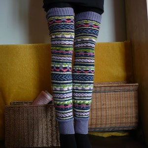 May include: A pair of colorful knitted leg warmers with a geometric pattern in shades of blue, green, pink, and purple. The leg warmers are displayed on a mannequin, showcasing their design and fit. The leg warmers are a cozy accessory.